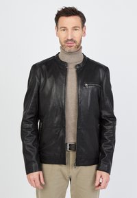 Man in een zwart leren jack over een beige turtleneck en beige broek, staande tegen een effen lichte achtergrond.