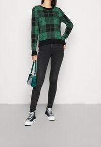 Pull à carreaux verts et noirs, jean noir slim, et baskets montantes noires. Tenant un petit sac à main texturé de couleur teal. Fond simple.