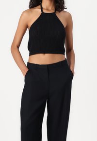 Zwarte gebreide haltercrop-top met geribbelde textuur, gecombineerd met zwarte high-waisted broek, handen in de zakken, neutrale achtergrond.