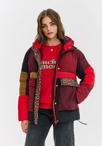 Žena s hnedými vlasmi po pleciach, oblečená v pestrofarebnej prešívanej bunde s leopardím vzorom, červenom mikinách a tmavých džínsoch, stojaca.