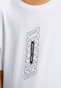 Biela tričko s čierne obrysy vertikálnym textom "DC SHOES" a menším čiernym vertikálnym prúžkom s nápisom "DCSHOECOUSA" v oblasti hrudníka.