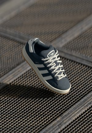 Mørkeblå Adidas Campus-sneaker med hvide striber og tykke snørebånd, hvilende på en tekstureret metalristeflade.
