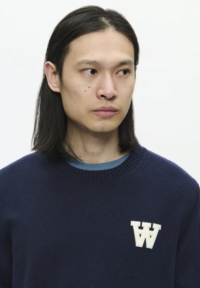 Navy blue knitted sweater with ribbed round neckline and white embroidered “W” logo on the left chest. Textured fabric with a soft appearance.