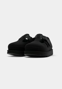 Paire de sandales noires à bout fermé avec semelles épaisses et brides ajustables, présentées sur un fond uni gris clair.