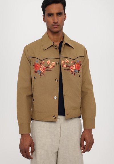 Brown cotton jacket featuring floral embroidery in red and blue, button closure, and a collared neck. Smooth texture, fitted design.