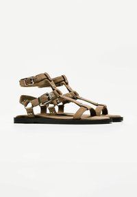Bruna mockaskinnsandaler med flera remmar, spännen och nitar. Platt sula med öppen tå-design och texturerad yta.