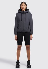 Veste légère grise avec capuche, fermeture éclair avant et poignets élastiques, associée à un short noir et des baskets noires. Détails de tissu texturé visibles.
