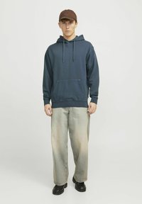 Sudadera azul marino con bolsillo tipo canguro, cordones y puños de canalé, combinada con jeans anchos de color claro y ligeramente descoloridos, y zapatos negros.