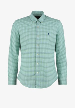 Homme portant une chemise à carreaux vert clair boutonnée avec manches retroussées, pantalon bleu marine foncé et baskets noires, debout les mains dans les poches.