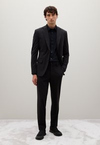 Traje negro a medida con corte ajustado, combinado con una camisa oscura y zapatos negros sin cordones. Tejido suave y líneas limpias. Fondo neutro.