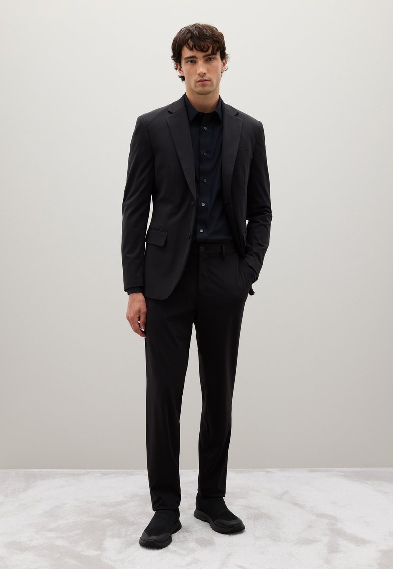 Traje negro a medida con corte ajustado, combinado con una camisa oscura y zapatos negros sin cordones. Tejido suave y líneas limpias. Fondo neutro.