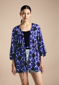 Kimono fluido blu con motivo, con cintura legata, maniche corte e scollatura a V profonda, indossato sopra un top nero e shorts in denim.