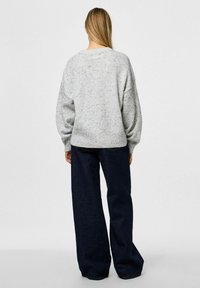 Femme aux longs cheveux blonds portant un pull gris clair à motifs tachetés et un jean large bleu foncé, debout de dos sur un fond uni.