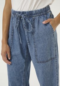 Gaiši zili džinsi ar elastīgu jostu, aukliņas detaļu un sānu kabatām. Audumam ir gluda tekstūra un maigs izbalējums.