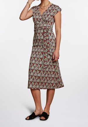 Kobieta w sukience do kolan z krótkim rękawem, z wielokolorowym abstrakcyjnym wzorem i paskiem przewiązanym w talii, czarnych sandałach wsuwanych oraz złotych naszyjnikach.