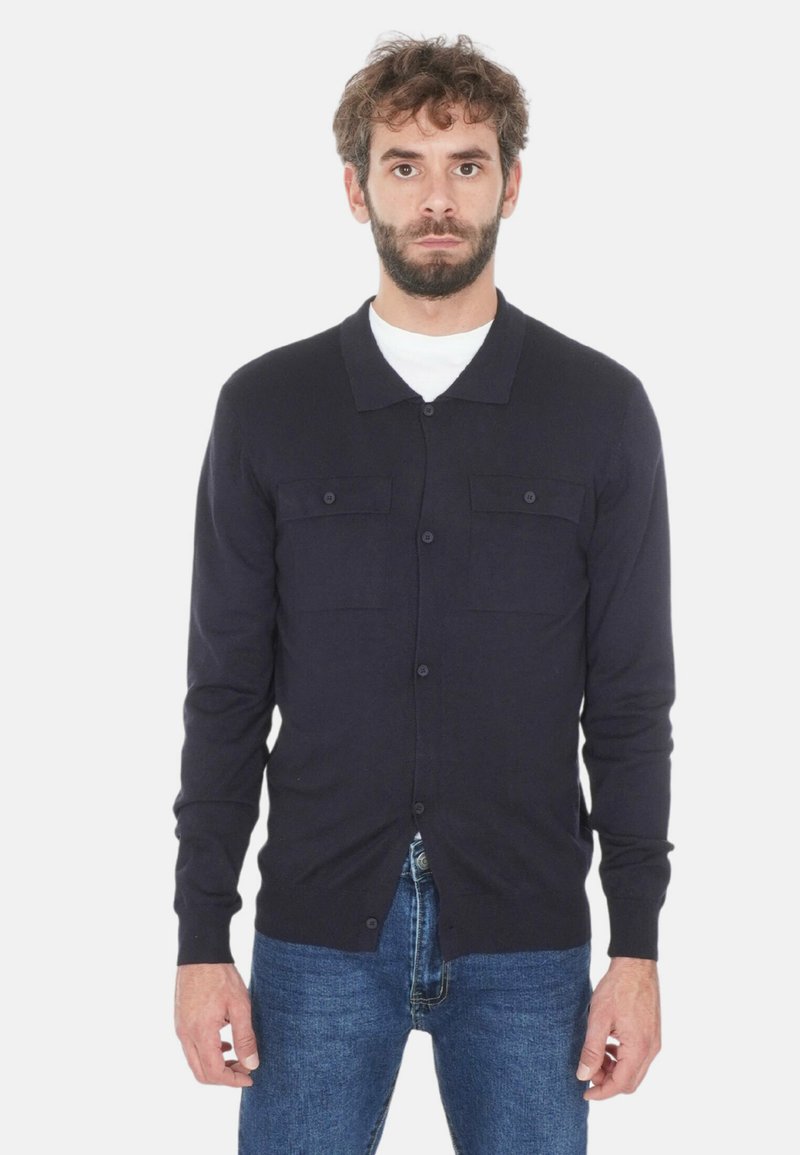 Uomo con capelli ricci e barba che indossa un cardigan buttoned blu scuro su una camicia bianca e jeans blu, in piedi davanti a uno sfondo neutro.