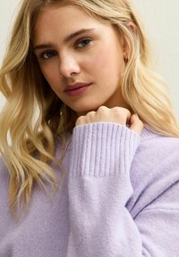 Blonde vrouw met golvend haar, gekleed in een lichtpaarse geribbelde trui, steunt met haar kin op haar vuist en kijkt zacht naar de camera.