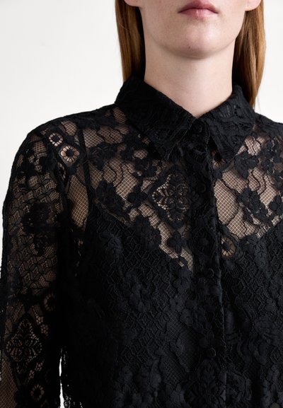 Blouse en dentelle noire avec col, présentant des motifs floraux complexes et une texture transparente, mettant en valeur des coutures détaillées et des accents de boutons.