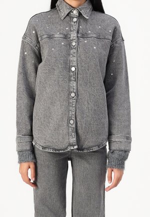 Femme portant une veste en jean grise avec des clous argentés et un jean gris assorti sur un fond blanc uni.