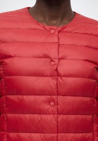 Personne portant une veste matelassée rouge avec des boutons ronds et un col sans revers.