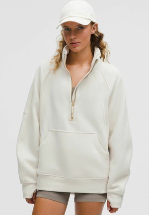 Jeune femme portant un sweatshirt oversized crème à zip quart, un short de vélo beige et une casquette crème assortie.