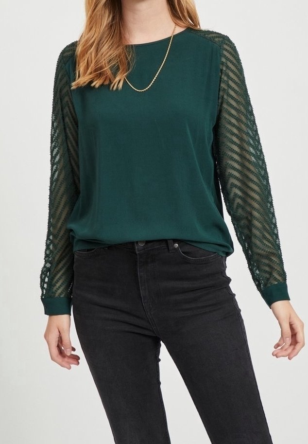 Grüne Bluse mit transparenten, gemusterten langen Ärmeln; runder Ausschnitt; kombiniert mit schwarzen Denim-Hosen. Akzente sind eine goldene Kettenhalskette.