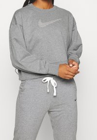 Szara bawełniana bluza z okrągłym dekoltem i długimi rękawami, z metalicznym logo Nike na piersi. W komplecie z szarymi spodniami dresowymi i białymi sznurkami.