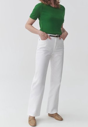 Personne portant un haut vert à manches courtes, un pantalon blanc taille haute avec une ceinture noire et des chaussures marron à enfiler, debout les mains dans les poches.