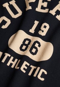 Zwart stof met beige tekst 'COLLEGE ATHLETIC', nummer '1986' in vetgedrukte vintage stijl. De stof is lichtelijk gekreukt.