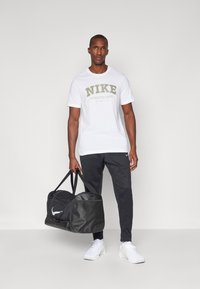 T-shirt bianco Nike con stampa verde e grigia, abbinato a pantaloni della tuta neri e scarpe da ginnastica bianche, mentre tiene una borsa a mano nera.