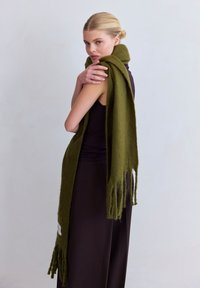Écharpe vert olive en tissu doux, avec des bords frangés, drapée sur l'épaule d'un mannequin portant une tenue sombre.