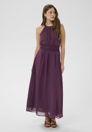 Femeie într-o rochie maxi mov închis fără mâneci, cu o centură de dantelă, păr lejer, bijuterii aurii și sandale maro cu vârf decupat, stând pe un fundal gri.