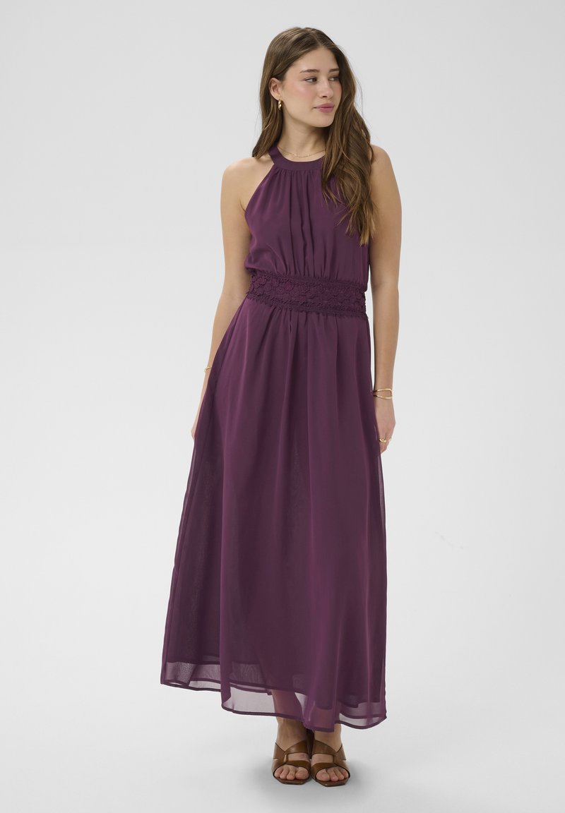 Femme portant une robe maxi sans manches de couleur violet foncé avec une ceinture en dentelle, cheveux lâchés, bijoux en or et sandales marron à bout ouvert, debout sur un fond gris.
