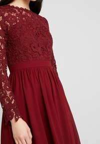 Burgundské šaty s krajkovým topem a dlouhými rukávy, s pevným pasem a splývavou sukní. Jemný květinový krajkový vzor na rukávech.
