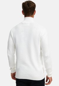 Mann sett bakfra iført en hvit teksturert strikkegenser med høy krage og svarte bukser mot en ensfarget bakgrunn.