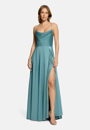 Femme en robe de soirée en satin couleur bleu sarcelle avec bretelles spaghetti et fente haute à la cuisse, portant des sandales argentées à talons hauts, se tenant devant un fond blanc.