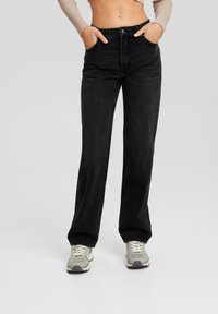 Personne portant un jean droit noir, un crop top côtelé beige et des baskets gris-blanc, debout les mains dans les poches.