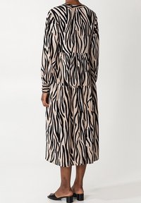 Pitkä, beige mekko mustilla seeprahaitansa, jossa on rento malli, pitkät hihat ja kerroksellinen hameosa. Yksinkertainen pääntie ja pehmeä kangas.