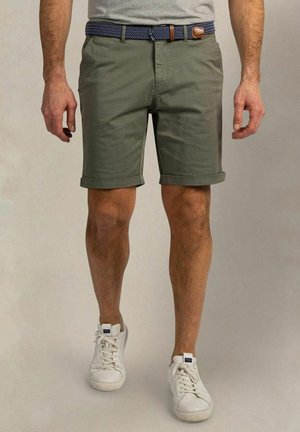 Man die groene opgerolde korte broek, witte sneakers, grijs T-shirt en een blauwe geweven riem met leren details draagt.