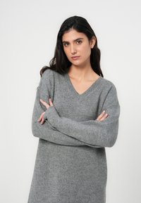 Γκρι πλεκτό πουλόβερ με V-λαιμόκοψη, μακριά μανίκια, άνετη εφαρμογή και υφή με ανάγλυφο φινίρισμα. Το ύφασμα φαίνεται μαλακό και ζεστό.