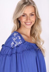 Blusa blu con maniche arricciate e ricamo floreale bianco sulla spalla. Tessuto morbido con un design ampio e fluido.