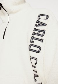 Pullover in pile bianco con colletto alto, caratterizzato dalla scritta ricamata in nero e blu "CARLO" lungo il lato. Superficie testurizzata.
