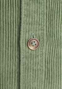 Vihreä corduroy-kangas, jossa on pystysuorat raidat, ja pyöreä ruskea nappi sekä siihen sopivaa vihreää ommelta sauman varrella.