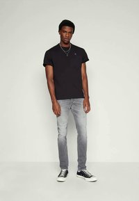 Mann steht vor einem neutralen Hintergrund und trägt eine schwarze Mütze, ein schwarzes T-Shirt, silberne Kreuzhalsketten, graue verwaschene Jeans und schwarze Low-Top-Sneaker.
