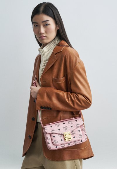 Kvinna i beige läderblazer och krämfärgad stickad tröja, håller en rosa designer crossbody-väska med guldspänne och svarta logotypmönster.