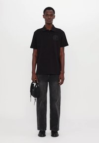 Svart polo skjorte med logo-patch, kombinert med mørkegrå rette jeans og svarte sko. Modellen holder en liten svart veske.