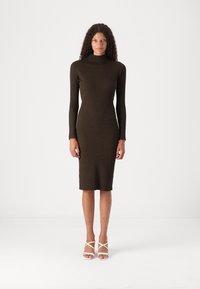 Robe en maille moulante marron avec des manches longues et un col montant, présentant une texture côtelée et une coupe au genou. Sandales blanches à brides.