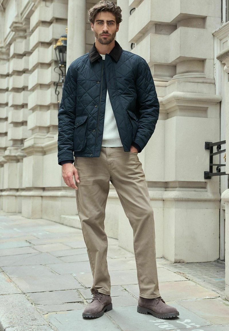Marineblau gesteppte Jacke mit braunem Kragen, weißem Pullover, beigen Hosen und braunen Schnürstiefeln, stehend auf einem Steinpflaster in einer städtischen Umgebung.
