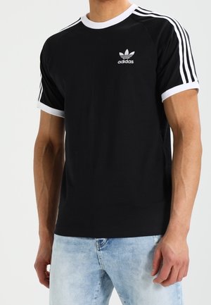 T-shirt noir Adidas avec bordure blanche et trois bandes blanches sur les manches, porté avec un jean bleu clair, montrant le haut du torse et les bras.