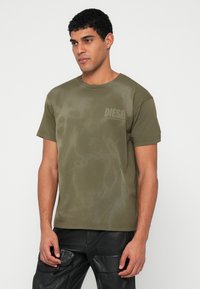 Λαδί βαμβακερό μπλουζάκι με στρογγυλή λαιμόκοψη, που διαθέτει ένα διακριτικό αφηρημένο σχέδιο και ένα μικρό, ανοιχτόχρωμο λογότυπο 'DIESEL' στο στήθος.